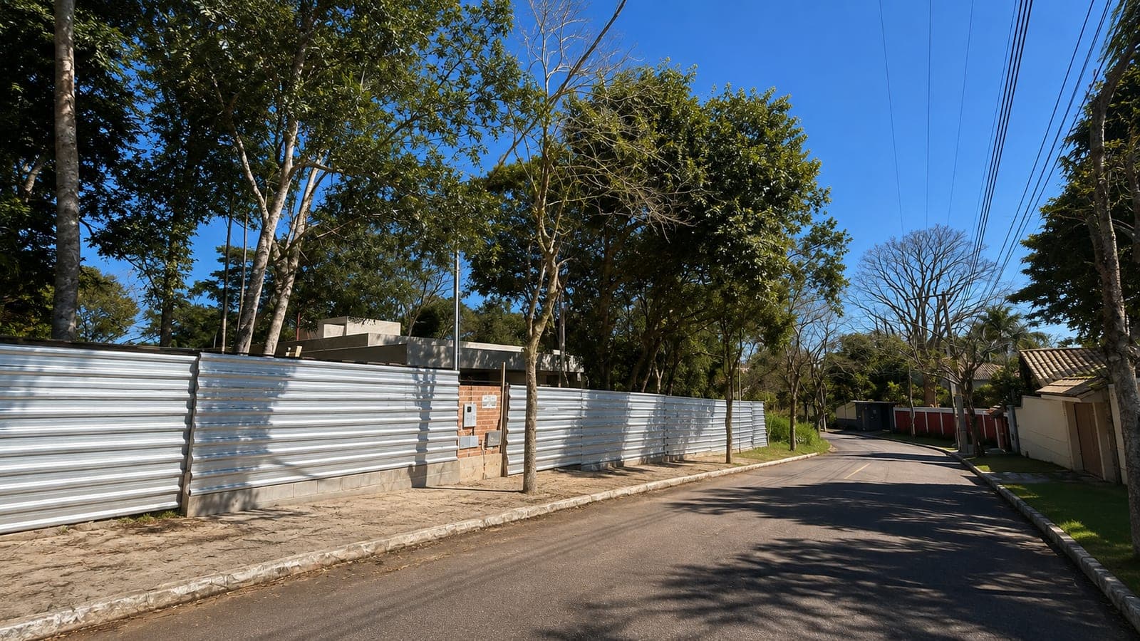 Rua arborizada em frente ao terreno, com o tapume da obra ao longo da calçada e a casa em construção entre as árvores
