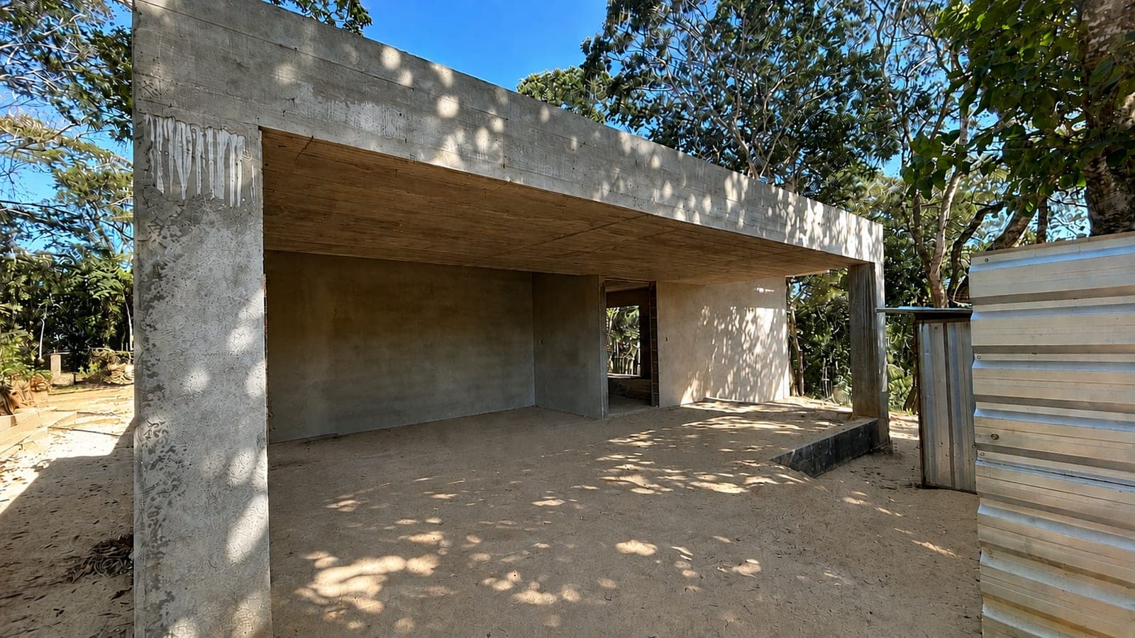 Garagem coberta por laje de concreto aparente, aberta para o terreno, com a entrada da sala ao fundo e a mata em volta