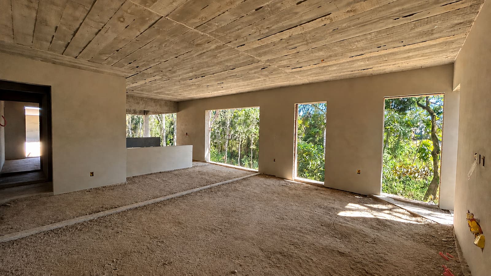Sala de estar e cozinha em obra, com laje de concreto aparente, paredes emboçadas, bancada da cozinha executada e quatro aberturas de piso ao teto voltadas para a mata
