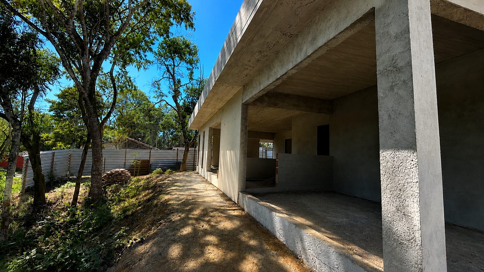 Lateral da casa em obra vista do jardim, com a laje avançando sobre os pilares de concreto e o caminho de terra ao lado das árvores