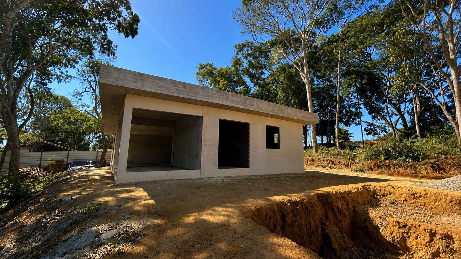 Fundos da casa principal em obra, com laje de cobertura em balanço, vãos abertos para o terreno em terraplenagem e a mata em volta