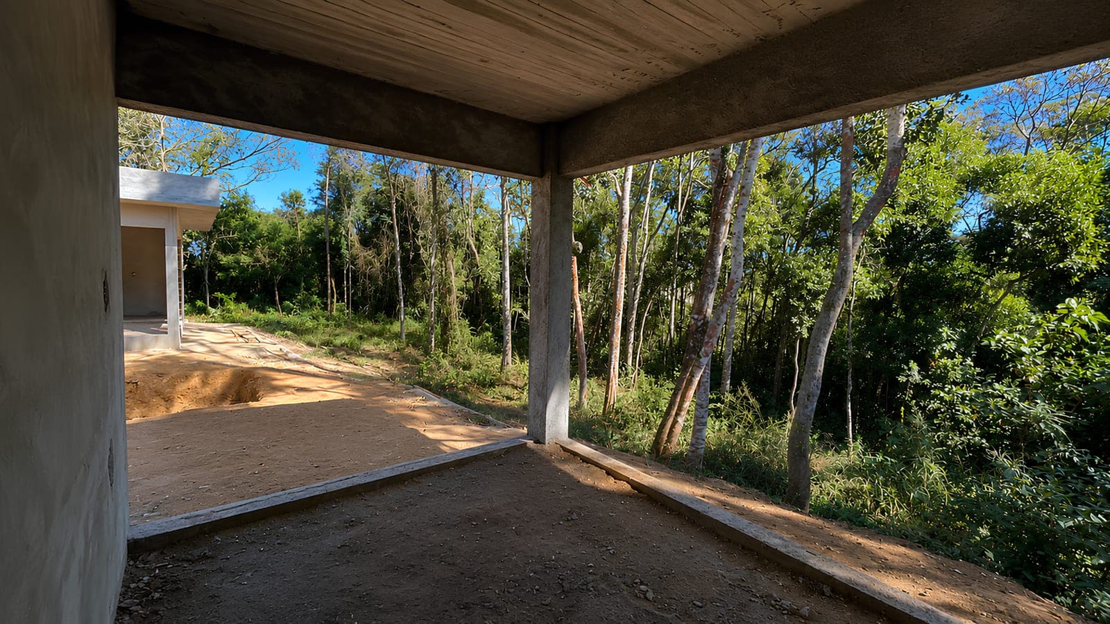 Varanda em obra, coberta por laje de concreto aparente apoiada em pilar, aberta para o bosque, com a outra edificação e o terreno em terraplenagem à esquerda