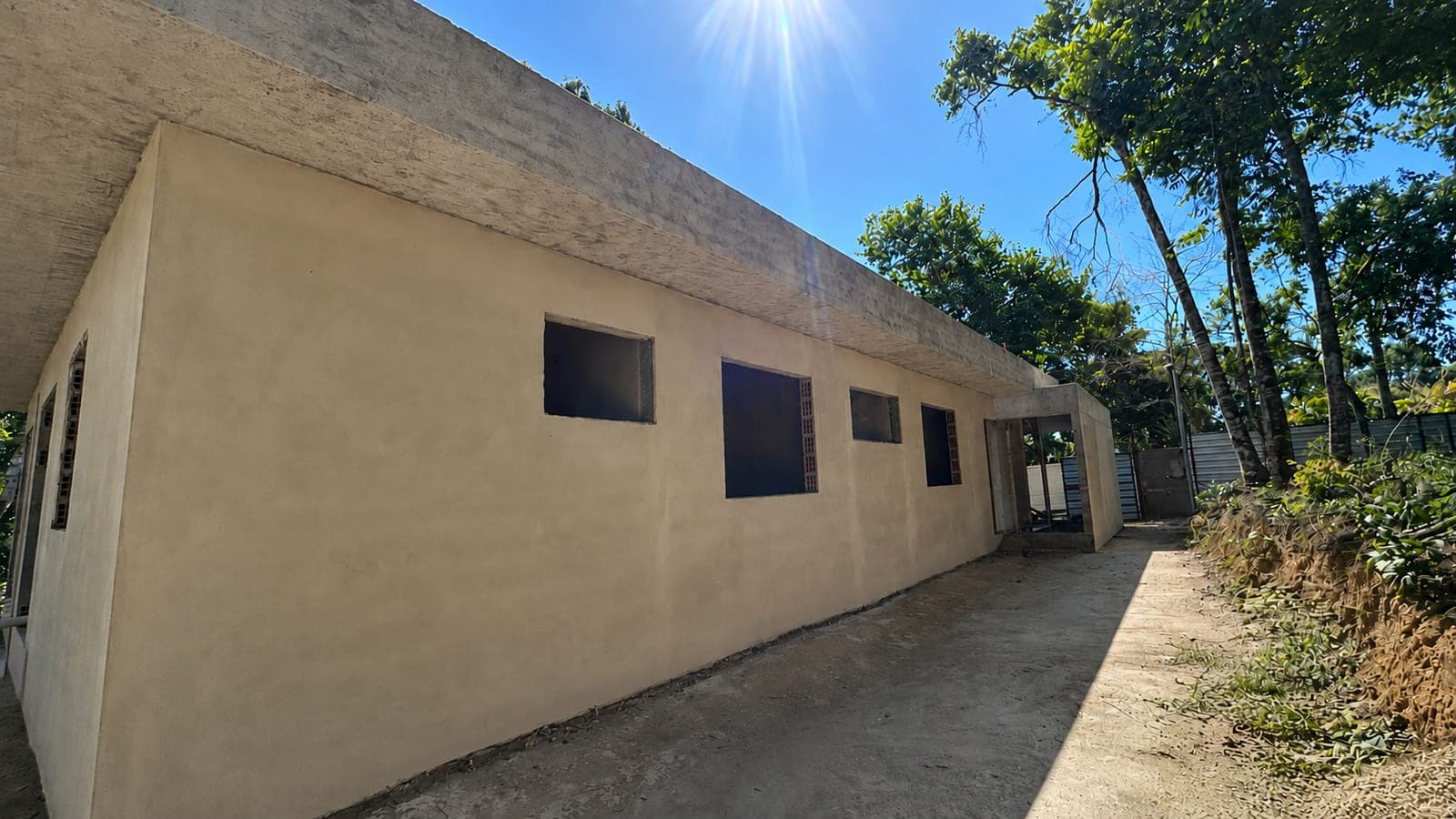 Lateral da casa em obra, com parede emboçada, vãos de janela sem esquadrias e o acesso lateral até os fundos do terreno